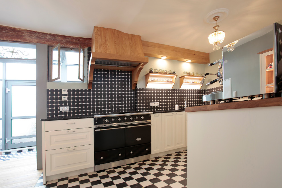 Example of a large country galley limestone floor and black floor open concept kitchen design in Nuremberg with a farmhouse sink, beaded inset cabinets, white cabinets, wood countertops, black backsplash, stone tile backsplash, black appliances, no island and brown countertops