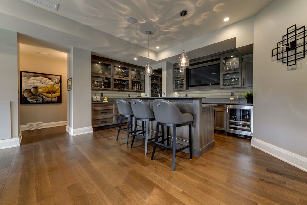This is an example of a large transitional l-shaped seated home bar in Edmonton with grey splashback, medium hardwood floors, glass tile splashback, an undermount sink, shaker cabinets, distressed cabinets, solid surface benchtops and brown floor.