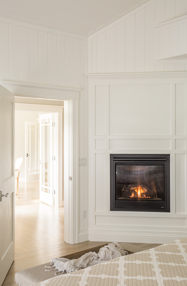 This is an example of an arts and crafts master bedroom in Portland Maine with white walls, light hardwood floors, a standard fireplace and a wood fireplace surround.