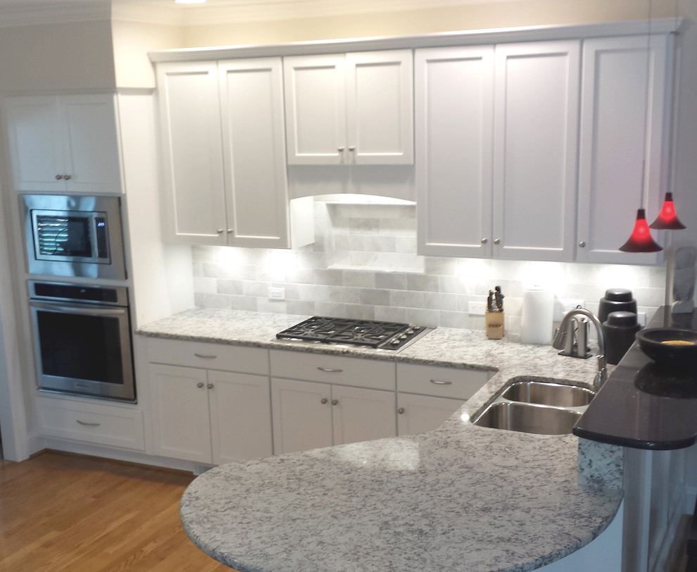 Example of a large transitional l-shaped light wood floor eat-in kitchen design with an undermount sink, recessed-panel cabinets, white cabinets, granite countertops, white backsplash, stone tile backsplash, white appliances and a peninsula