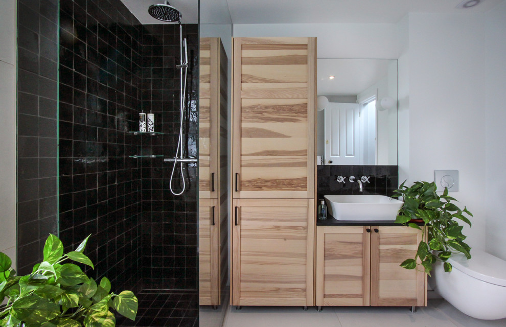 This is an example of a mid-sized contemporary kids bathroom in London with shaker cabinets, light wood cabinets, a drop-in tub, an open shower, a wall-mount toilet, black tile, terra-cotta tile, white walls, porcelain floors, a console sink, white floor and an open shower.