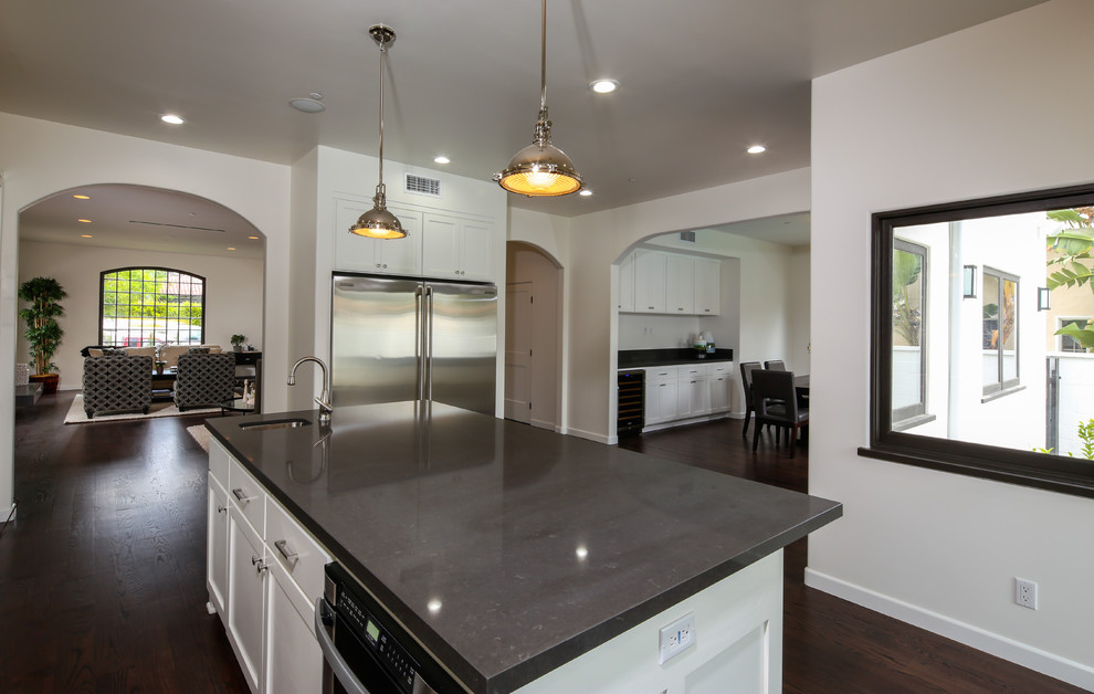 Large transitional l-shaped kitchen in Los Angeles with a farmhouse sink, shaker cabinets, white cabinets, quartz benchtops, grey splashback, stone tile splashback, stainless steel appliances, dark hardwood floors and with island.