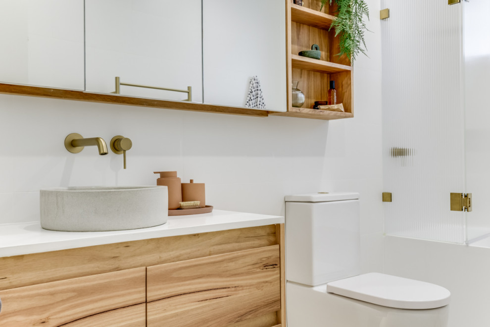 Inspiration for a small modern family bathroom in Sydney with flat-panel cabinets, light wood cabinets, a corner bath, a shower/bath combination, a two-piece toilet, white tiles, white walls, a vessel sink, grey floors, a hinged door, white worktops, a wall niche, a single sink and a floating vanity unit.