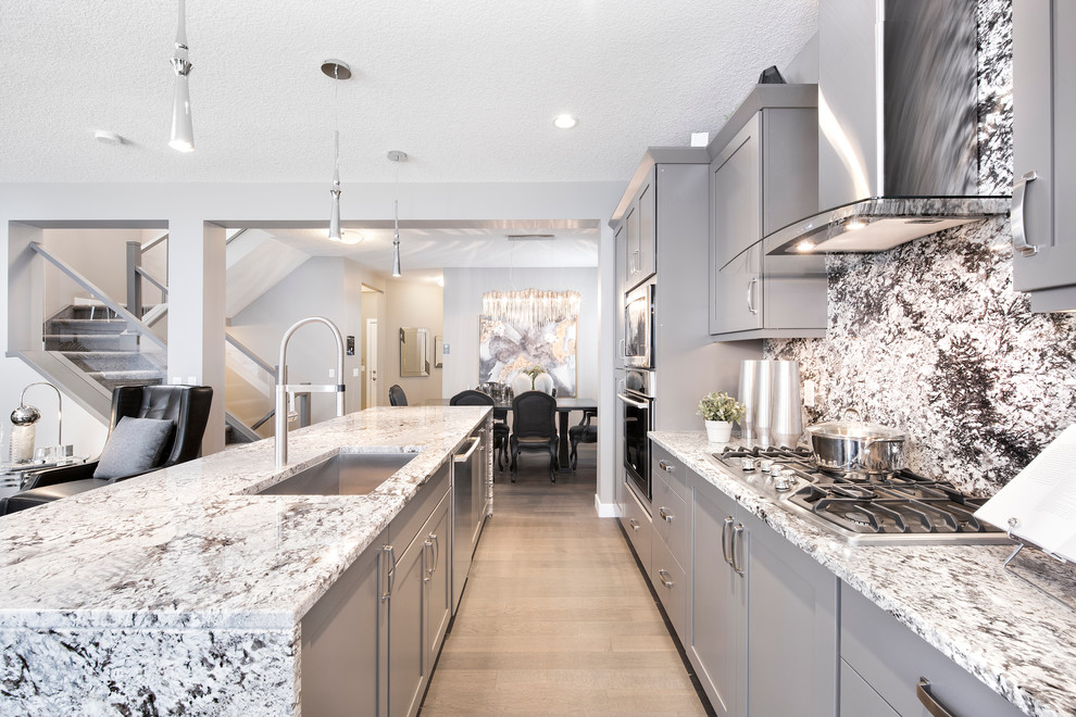 Mid-sized contemporary galley open plan kitchen in Edmonton with an undermount sink, flat-panel cabinets, grey cabinets, granite benchtops, grey splashback, stone slab splashback, stainless steel appliances, light hardwood floors and with island.