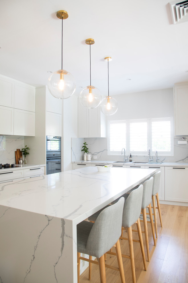 This is an example of an expansive transitional l-shaped open plan kitchen in Sydney with a double-bowl sink, shaker cabinets, white cabinets, quartz benchtops, white splashback, marble splashback, stainless steel appliances, light hardwood floors, with island, beige floor and white benchtop.
