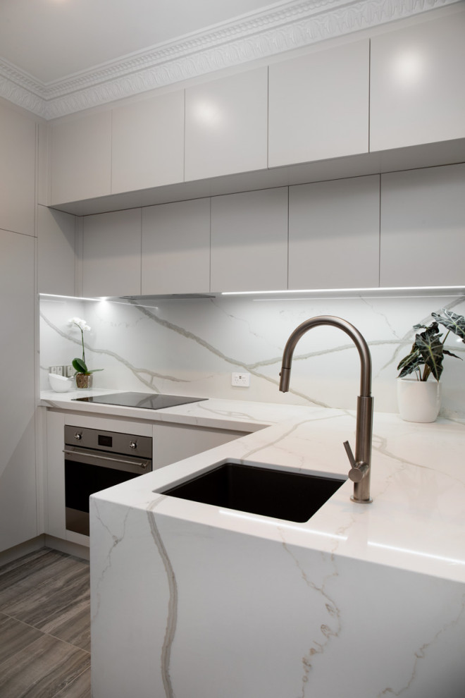 This is an example of a small contemporary l-shaped open plan kitchen in Brisbane with an undermount sink, flat-panel cabinets, beige cabinets, quartz benchtops, white splashback, engineered quartz splashback, stainless steel appliances, porcelain floors, no island, grey floor, white benchtop and recessed.