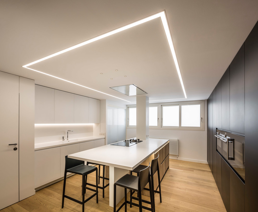 Design ideas for a large contemporary u-shaped open plan kitchen in Valencia with an integrated sink, flat-panel cabinets, white cabinets, quartz benchtops, white splashback, black appliances, medium hardwood floors, with island, brown floor and white benchtop.