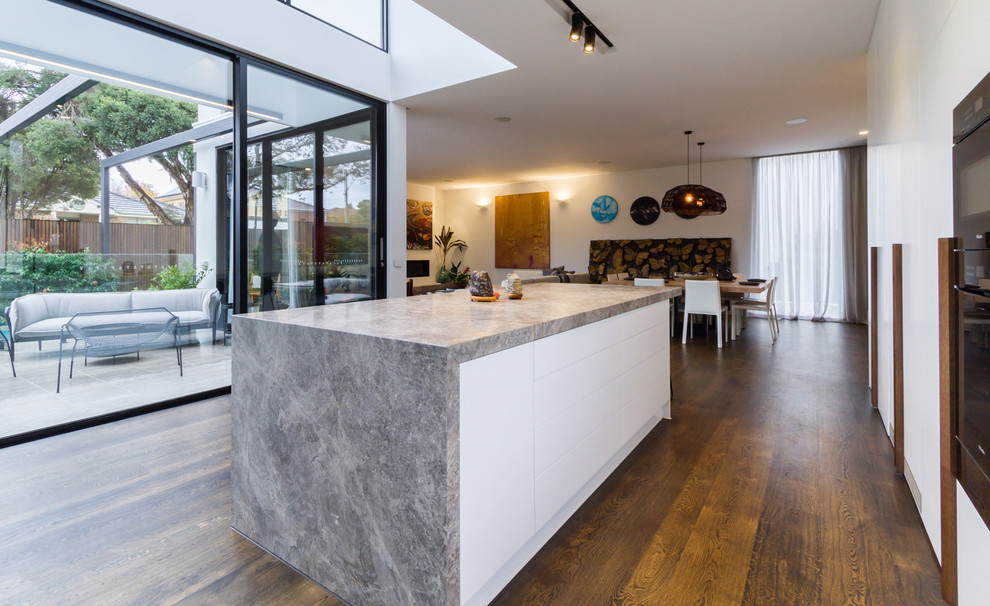 Photo of a large modern l-shaped open plan kitchen in Melbourne with an undermount sink, flat-panel cabinets, white cabinets, grey splashback, marble splashback, black appliances, medium hardwood floors, with island, brown floor, grey benchtop and limestone benchtops.