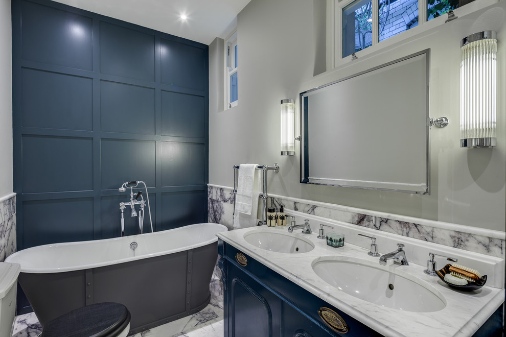 Mid-sized traditional master bathroom in London with an undermount sink, blue cabinets, marble benchtops, a freestanding tub, a two-piece toilet, white tile, stone tile, grey walls, marble floors and recessed-panel cabinets.