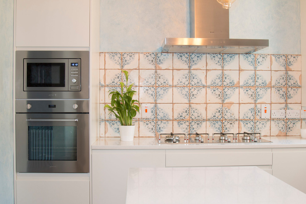Small bohemian l-shaped open plan kitchen with a built-in sink, flat-panel cabinets, beige cabinets, quartz worktops, beige splashback, cement tile splashback, stainless steel appliances, laminate floors, an island and brown floors.