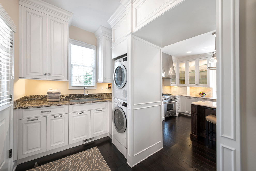 Design ideas for a traditional l-shaped laundry room in Detroit with an undermount sink, raised-panel cabinets, white cabinets, beige walls, dark hardwood floors and a stacked washer and dryer.