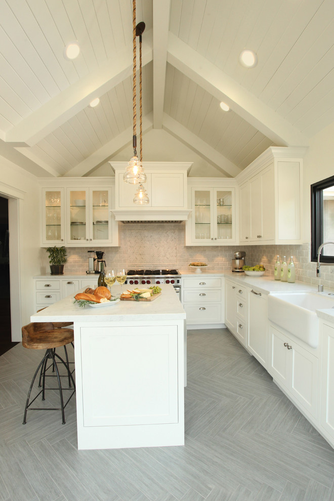 Large transitional l-shaped kitchen in Los Angeles with a farmhouse sink, shaker cabinets, stone tile splashback, stainless steel appliances, ceramic floors, grey floor, white splashback, with island, grey benchtop and white cabinets.
