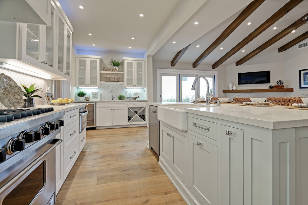 Design ideas for a beach style u-shaped open plan kitchen in Los Angeles with a farmhouse sink, shaker cabinets, white cabinets, white splashback, subway tile splashback, stainless steel appliances, light hardwood floors and with island.