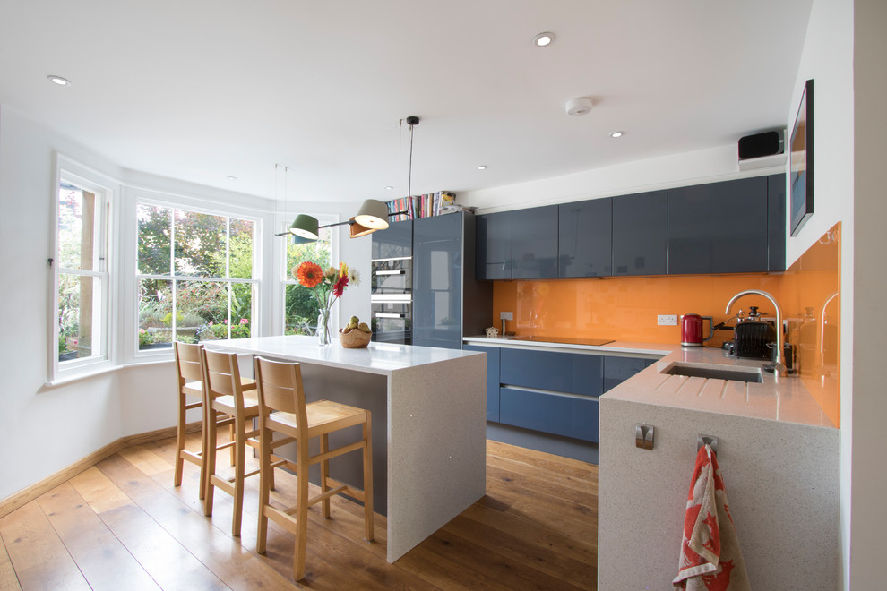 Design ideas for a contemporary l-shaped kitchen in Other with an undermount sink, flat-panel cabinets, blue cabinets, orange splashback, glass sheet splashback, black appliances, medium hardwood floors, with island and white benchtop.