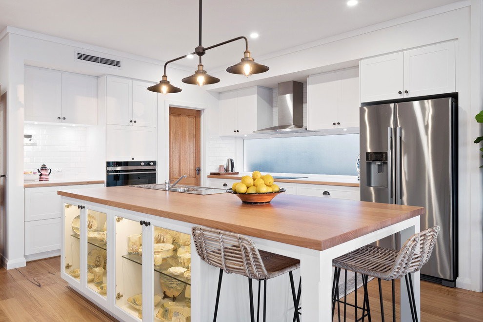 Mid-sized beach style l-shaped kitchen in Perth with a drop-in sink, recessed-panel cabinets, white cabinets, wood benchtops, white splashback, subway tile splashback, stainless steel appliances, medium hardwood floors, with island, brown floor and brown benchtop.
