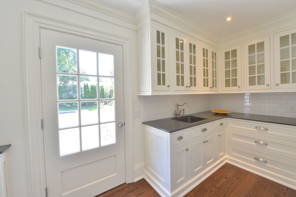 Mid-sized transitional l-shaped utility room in New York with an undermount sink, glass-front cabinets, white cabinets, quartz benchtops, white walls, medium hardwood floors and brown floor.