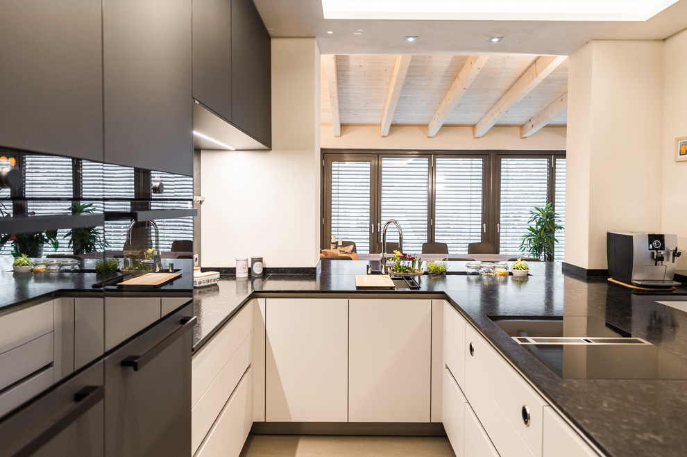 This is an example of a mid-sized contemporary u-shaped open plan kitchen in Leipzig with an undermount sink, flat-panel cabinets, grey cabinets, granite benchtops, grey splashback, stone slab splashback, black appliances, a peninsula, grey floor and grey benchtop.