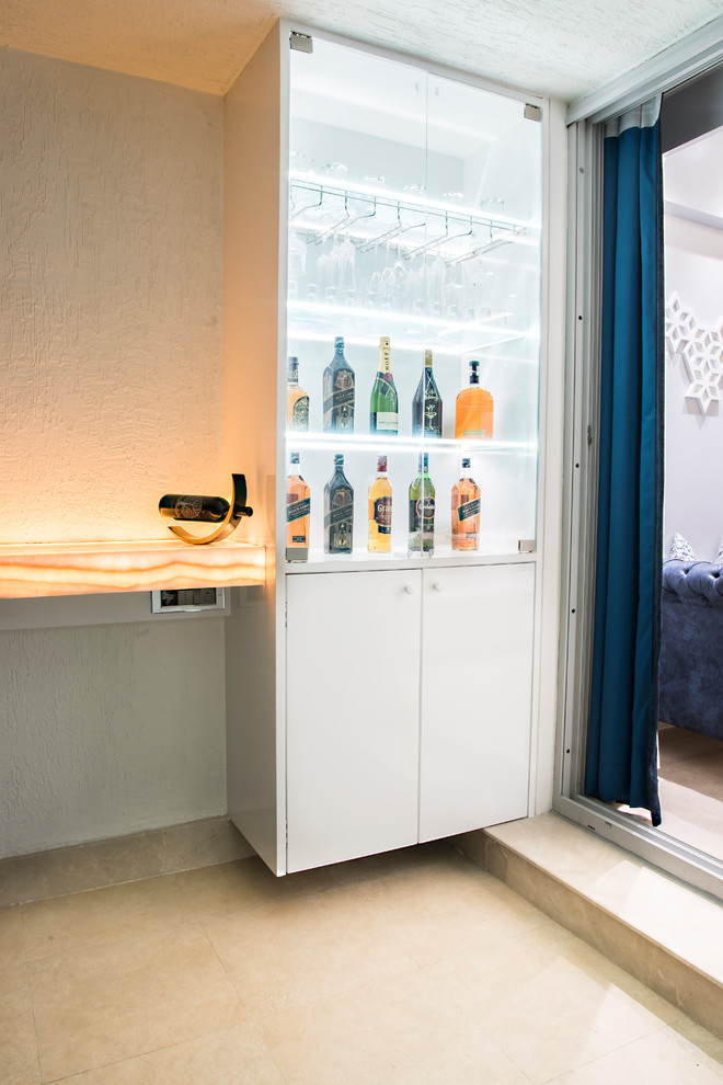 This is an example of a contemporary home bar in Mumbai with raised-panel cabinets, white cabinets, onyx benchtops, white splashback, ceramic floors, beige floor and beige benchtop.