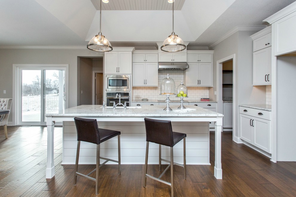 Mid-sized transitional single-wall medium tone wood floor and beige floor open concept kitchen photo with a farmhouse sink, recessed-panel cabinets, white cabinets, granite countertops, white backsplash, ceramic backsplash, stainless steel appliances and an island