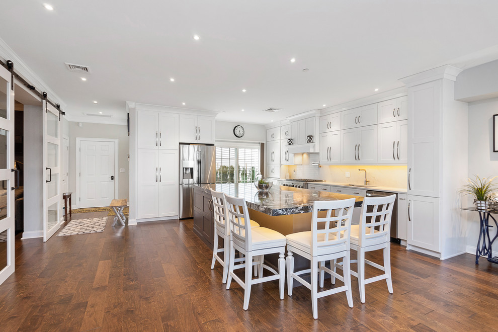 Example of a mid-sized transitional l-shaped medium tone wood floor and brown floor open concept kitchen design in Boston with an undermount sink, shaker cabinets, white cabinets, quartz countertops, white backsplash, stainless steel appliances, an island and white countertops