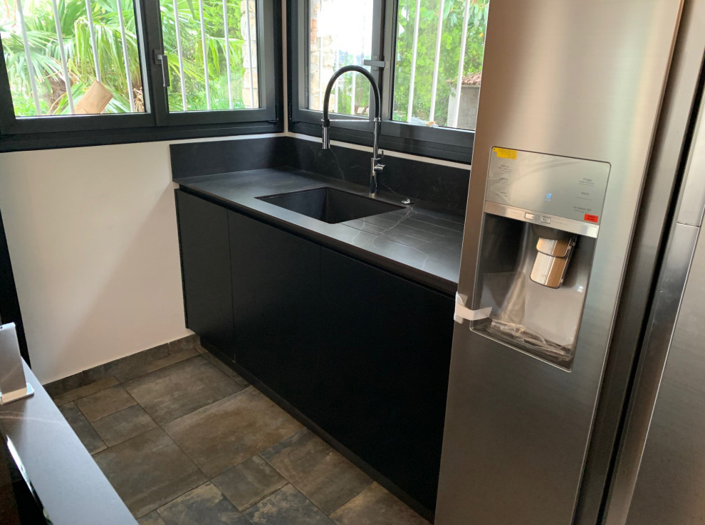 Mid-sized contemporary single-wall open plan kitchen in Nice with a single-bowl sink, flat-panel cabinets, black cabinets, black splashback, engineered quartz splashback, stainless steel appliances, ceramic floors, a peninsula, multi-coloured floor, grey benchtop and recessed.