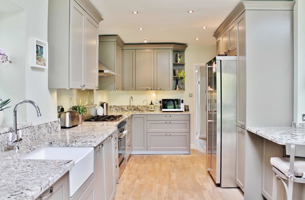 Photo of a mid-sized transitional l-shaped kitchen in London with a farmhouse sink, recessed-panel cabinets, grey cabinets, stainless steel appliances, a peninsula and beige floor.