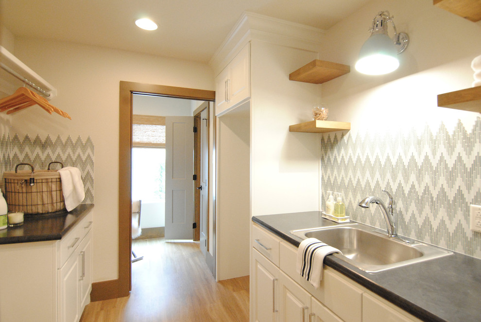 Photo of a large eclectic galley utility room with a drop-in sink, recessed-panel cabinets, white cabinets, laminate benchtops, white walls, vinyl floors, a side-by-side washer and dryer and brown floor.