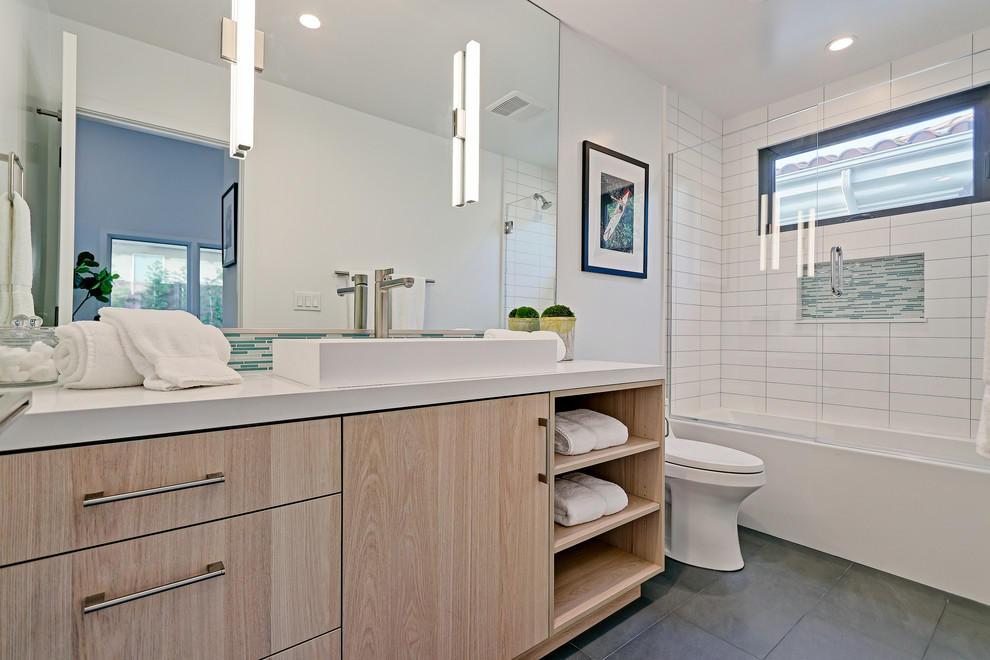 Inspiration for a mid-sized contemporary bathroom in Los Angeles with flat-panel cabinets, light wood cabinets, an alcove tub, a shower/bathtub combo, a one-piece toilet, white tile, subway tile, white walls, porcelain floors, a vessel sink, solid surface benchtops, grey floor and a hinged shower door.