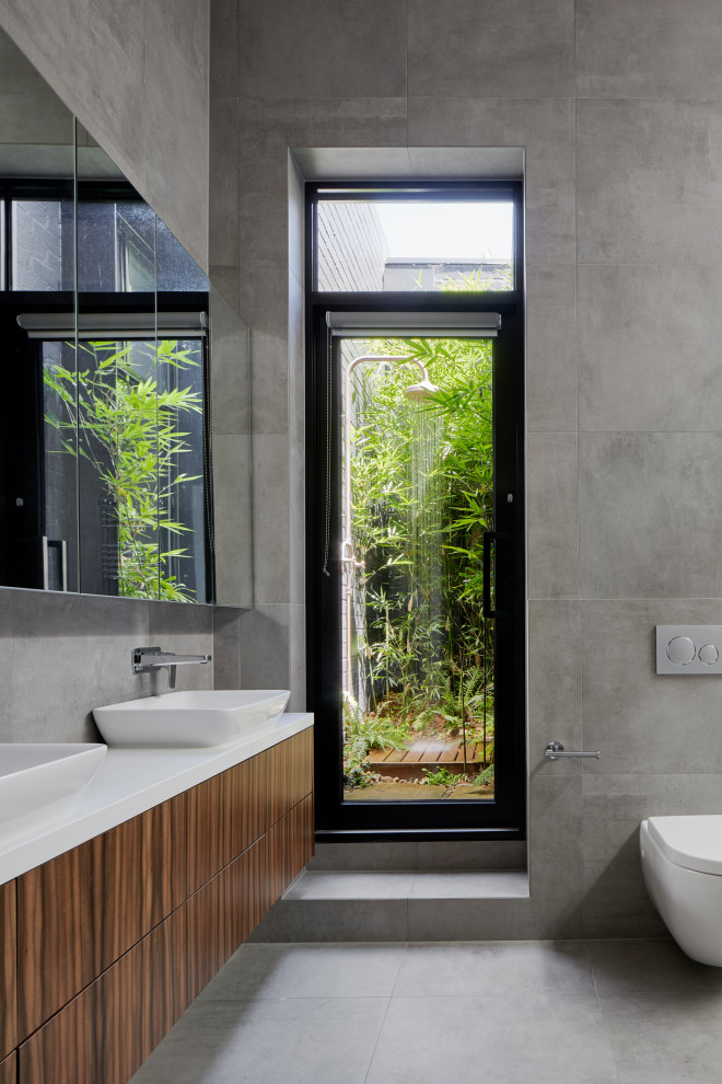 Large contemporary master wet room bathroom in Melbourne with medium wood cabinets, a wall-mount toilet, gray tile, porcelain tile, grey floor, white benchtops, a freestanding tub, grey walls, porcelain floors, a drop-in sink, engineered quartz benchtops, an open shower and flat-panel cabinets.