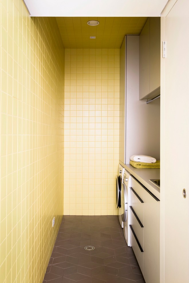 Photo of a small contemporary galley dedicated laundry room in Sydney with a drop-in sink, flat-panel cabinets, grey cabinets, laminate benchtops, yellow walls, porcelain floors and a side-by-side washer and dryer.