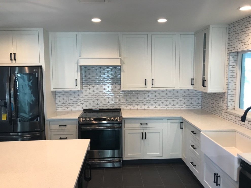Photo of a mid-sized midcentury l-shaped kitchen in Tampa with a farmhouse sink, shaker cabinets, white cabinets, quartz benchtops, white splashback, porcelain splashback, stainless steel appliances, porcelain floors, with island, grey floor and white benchtop.