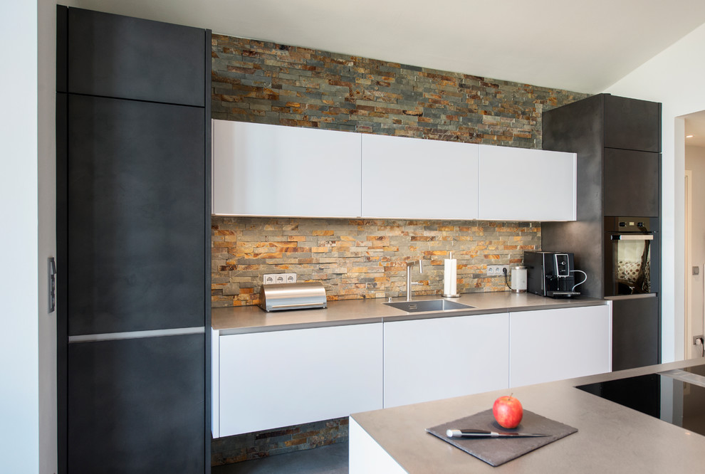 This is an example of a mid-sized contemporary open plan kitchen in Nuremberg with an integrated sink, flat-panel cabinets, multi-coloured splashback, stone tile splashback, black appliances, concrete floors, with island, grey floor and grey benchtop.
