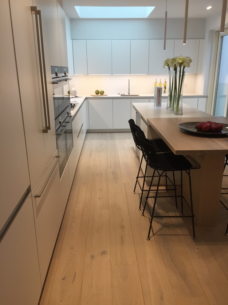 This is an example of a large contemporary l-shaped kitchen/diner in Buckinghamshire with an integrated sink, flat-panel cabinets, white cabinets, composite countertops, white splashback, glass sheet splashback, stainless steel appliances, medium hardwood flooring and an island.