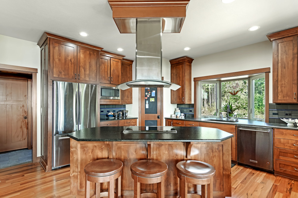 Example of a transitional l-shaped medium tone wood floor and brown floor kitchen design in Seattle with an undermount sink, shaker cabinets, medium tone wood cabinets, gray backsplash, stainless steel appliances, an island and black countertops