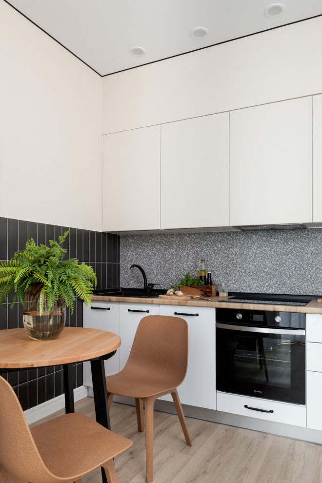 Contemporary single-wall kitchen in Other with flat-panel cabinets, white cabinets, grey splashback, black appliances, light hardwood floors, no island, beige floor and brown benchtop.