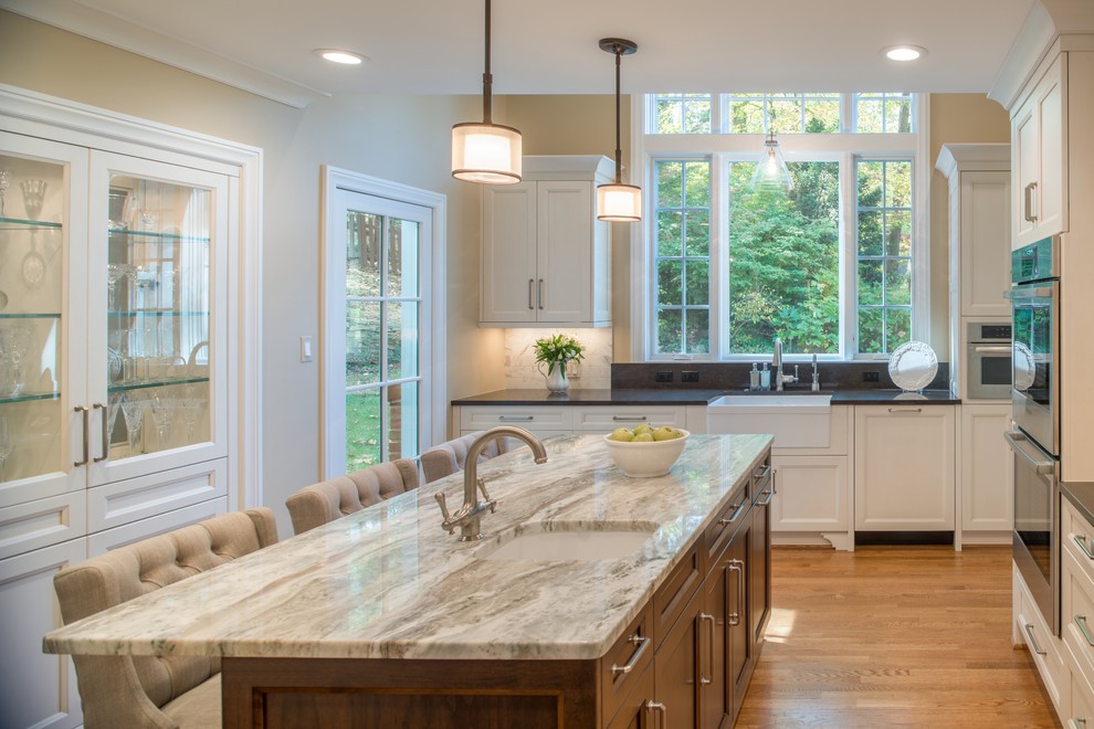 Example of a mid-sized transitional u-shaped medium tone wood floor eat-in kitchen design in DC Metro with a farmhouse sink, shaker cabinets, white cabinets, quartzite countertops, gray backsplash, subway tile backsplash, stainless steel appliances and an island