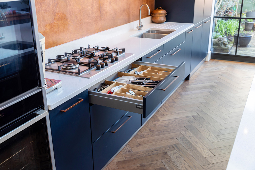 Inspiration for a large contemporary single-wall eat-in kitchen in London with an undermount sink, flat-panel cabinets, blue cabinets, quartzite benchtops, metallic splashback, black appliances, medium hardwood floors, with island, brown floor and white benchtop.