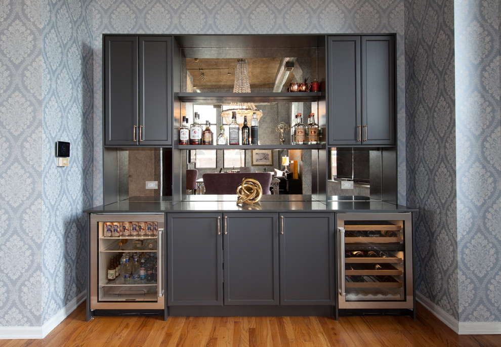 This is an example of a mid-sized eclectic single-wall home bar in Chicago with recessed-panel cabinets, grey cabinets, quartz benchtops, grey splashback, mirror splashback, light hardwood floors, brown floor and grey benchtop.