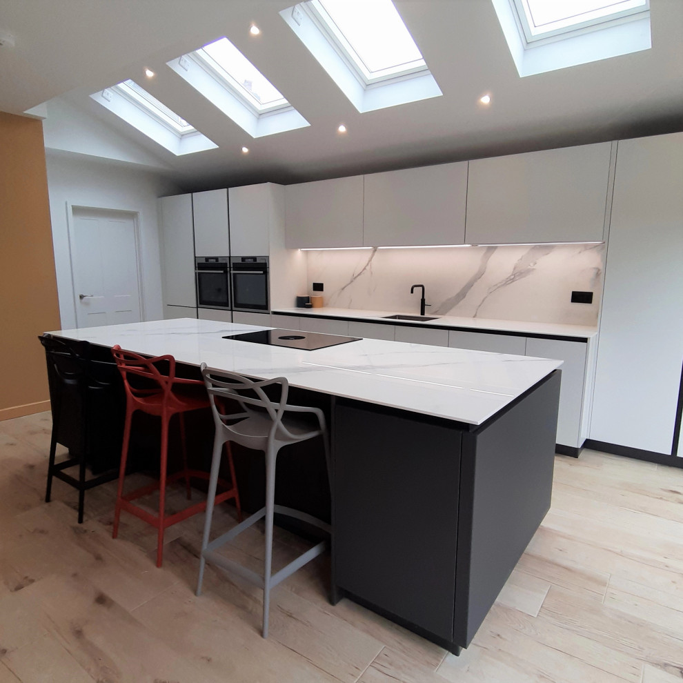 Large contemporary single-wall open plan kitchen in Hertfordshire with a single-bowl sink, flat-panel cabinets, white cabinets, quartzite benchtops, white splashback, stainless steel appliances, light hardwood floors, with island, white benchtop and vaulted.