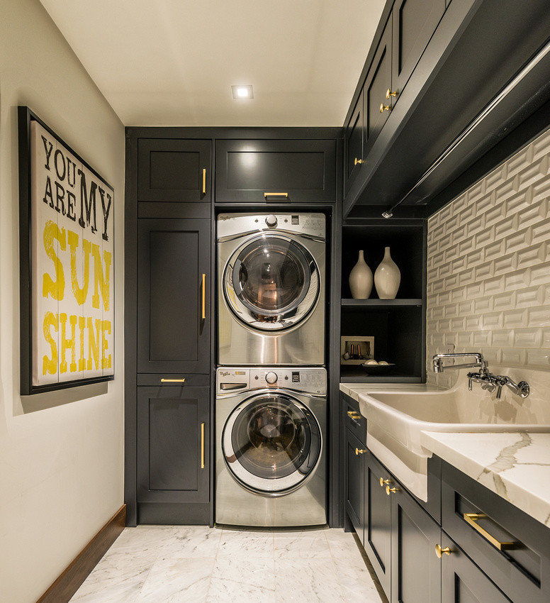 Inspiration for a mid-sized transitional l-shaped laundry room with a farmhouse sink, shaker cabinets, marble benchtops, white walls, marble floors, a stacked washer and dryer, white floor and black cabinets.