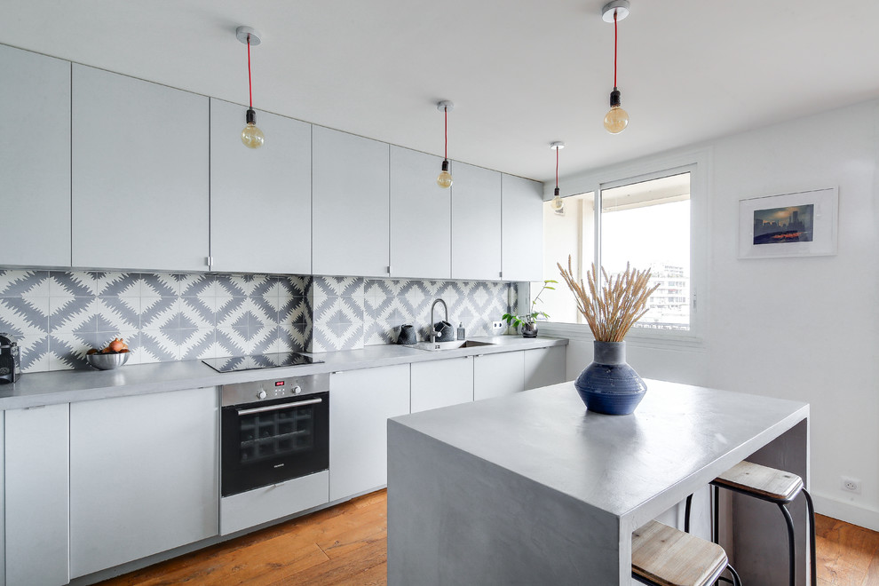 Inspiration pour une grande cuisine américaine design en L avec un évier encastré, un placard à porte affleurante, des portes de placard grises, un plan de travail en béton, une crédence grise, une crédence en carreau de ciment, un électroménager en acier inoxydable, un sol en bois brun, îlot, un sol marron et un plan de travail gris.