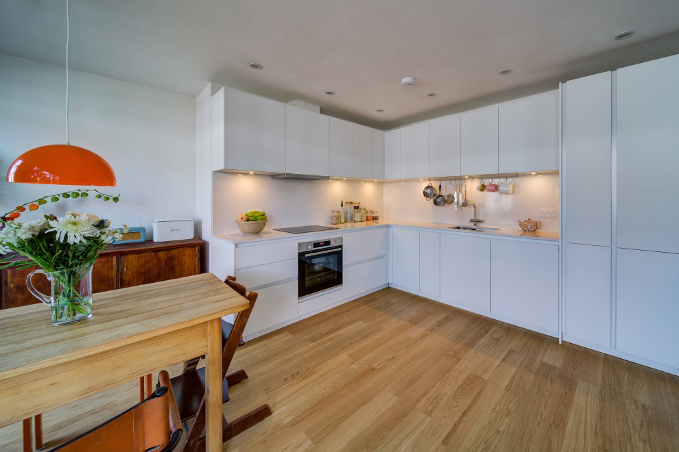 This is an example of a mid-sized contemporary l-shaped eat-in kitchen in London with a double-bowl sink, flat-panel cabinets, white cabinets, quartzite benchtops, white splashback, black appliances, light hardwood floors, no island, brown floor and white benchtop.
