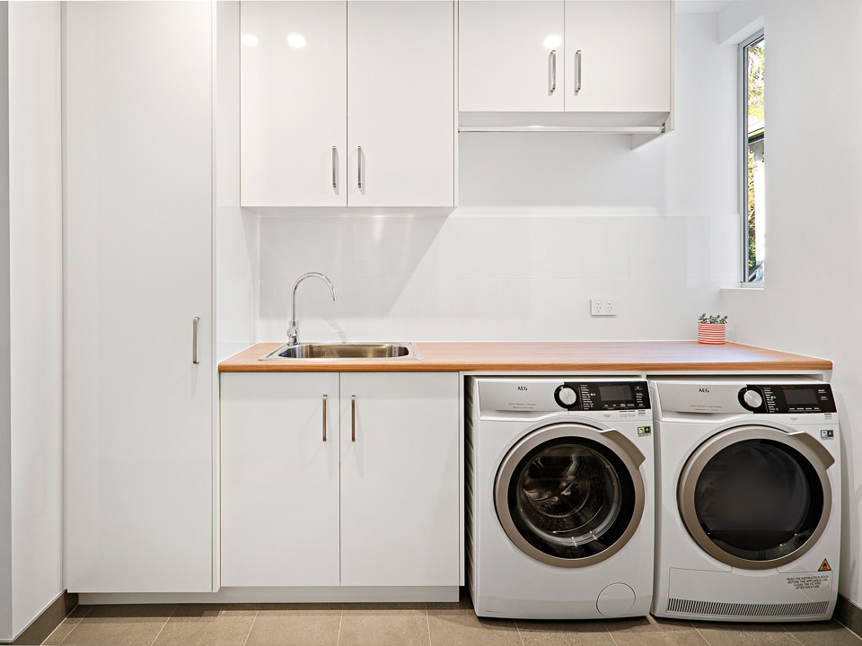 Small modern single-wall dedicated laundry room in Brisbane with a single-bowl sink, flat-panel cabinets, white cabinets, laminate benchtops, white walls, porcelain floors, a side-by-side washer and dryer, grey floor and brown benchtop.