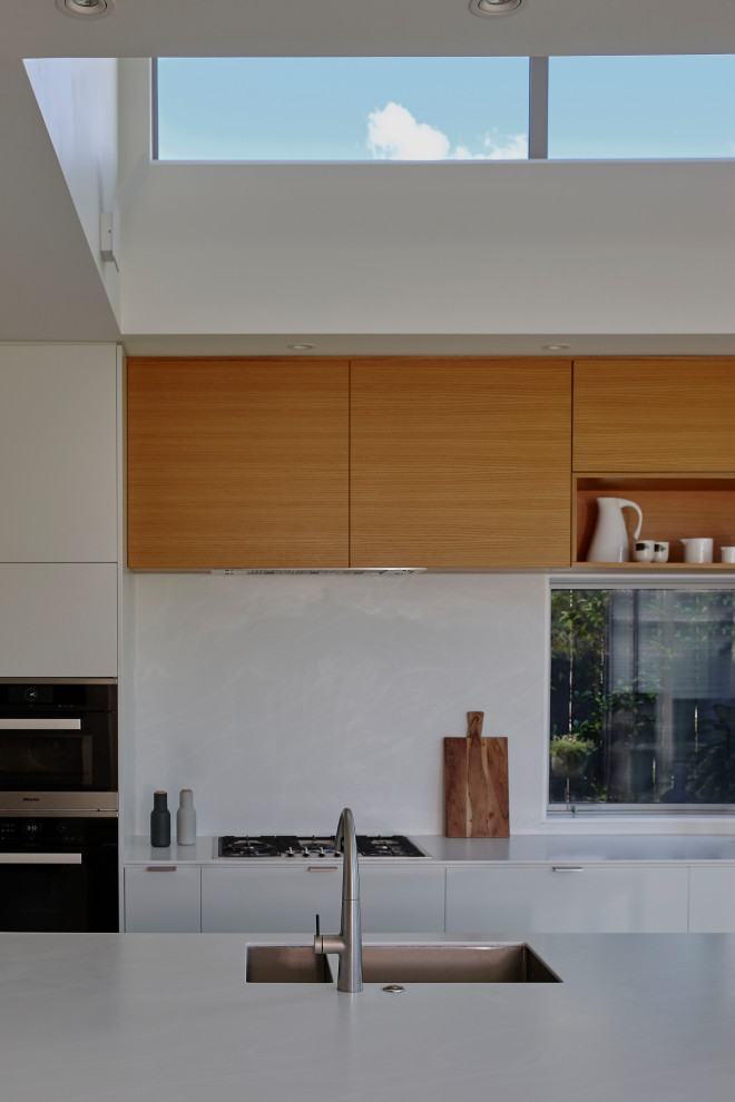 Photo of a medium sized contemporary galley kitchen pantry in Auckland with an integrated sink, flat-panel cabinets, light wood cabinets, composite countertops, white splashback, engineered quartz splashback, stainless steel appliances, concrete flooring, an island, grey floors and white worktops.