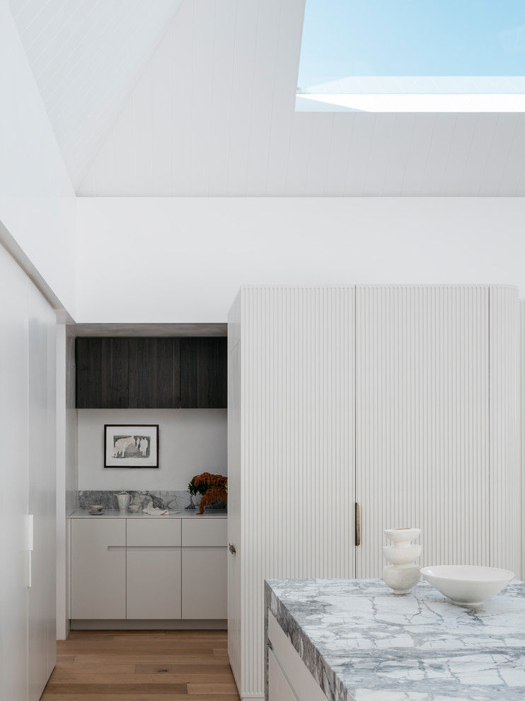 Large contemporary single-wall eat-in kitchen in Sydney with an undermount sink, flat-panel cabinets, white cabinets, granite benchtops, white splashback, white appliances, light hardwood floors, with island, brown floor and white benchtop.