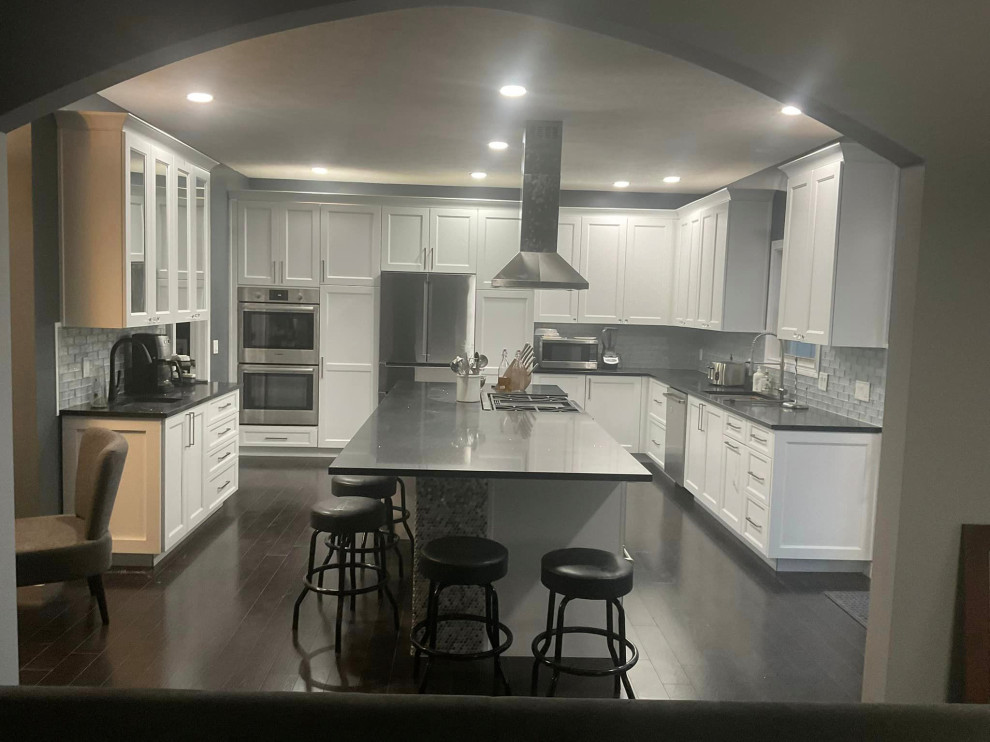 Photo of a large contemporary kitchen in Other with a single-bowl sink, recessed-panel cabinets, white cabinets, quartz benchtops, white splashback, glass tile splashback, stainless steel appliances, dark hardwood floors, with island and black benchtop.