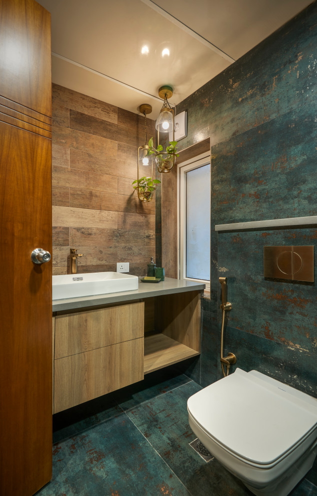 Bild på ett industriellt grå grått toalett, med släta luckor, skåp i ljust trä, en vägghängd toalettstol, flerfärgad kakel, ett fristående handfat och turkost golv
