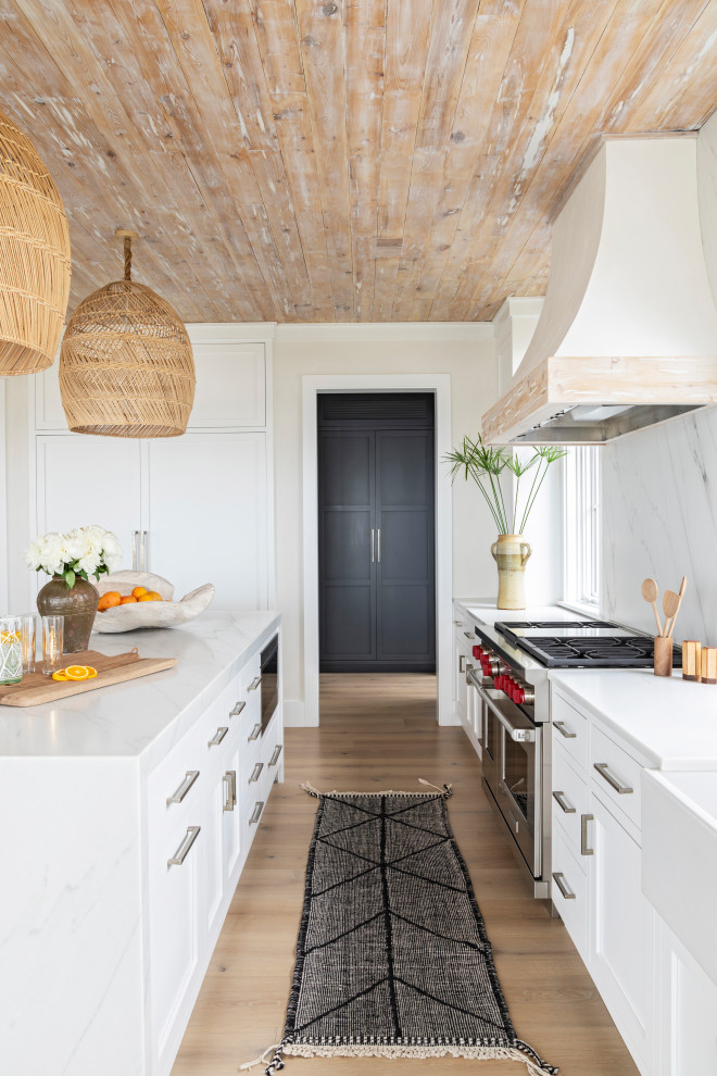 Inspiration for an expansive coastal single-wall open plan kitchen in Charleston with a belfast sink, recessed-panel cabinets, white cabinets, marble worktops, white splashback, marble splashback, integrated appliances, light hardwood flooring, an island, beige floors, white worktops and a wood ceiling.
