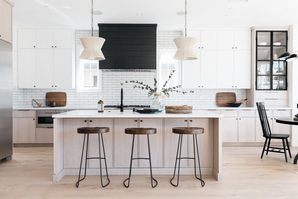 This is an example of a large country galley open plan kitchen in Los Angeles with flat-panel cabinets, white cabinets, white splashback, subway tile splashback, stainless steel appliances, light hardwood floors, with island, beige floor and white benchtop.