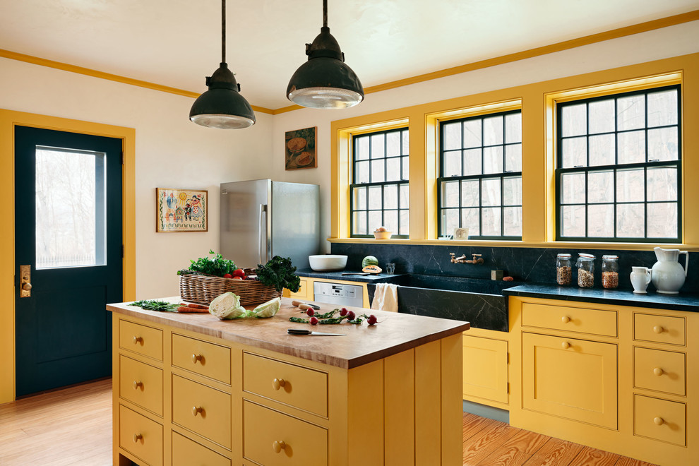 Photo of a mid-sized country kitchen in New York with a farmhouse sink, yellow cabinets, soapstone benchtops, black splashback, stone slab splashback, stainless steel appliances, with island, black benchtop, beaded inset cabinets and light hardwood floors.
